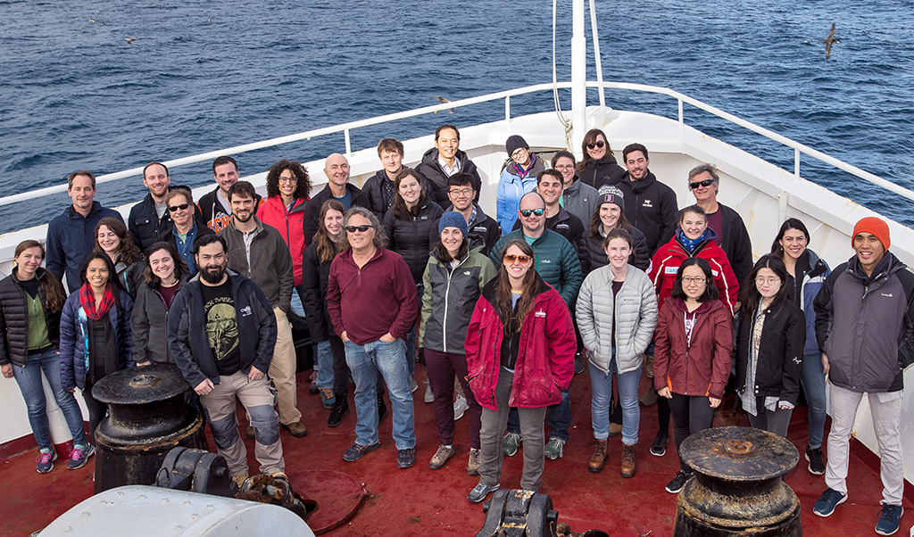 34 people in cold-weather gear posing in group at front of ship in ocean.